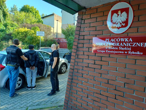  Funkcjonariusze prowadzą osobę zatrzymaną do samochodu, na budynku napis: Placówka Straży Granicznej w Rudzie Śląskiej Grupa Zamiejscowa w Rybniku.