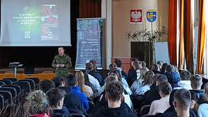 Od lewej ekran z wyświetloną prezentacją, funkcjonariusz SG, rollup SG w Opolu, na ścianie Godło Polski i herb. Na krzesłach siedzą uczniowie. Zdjęcie wykonane z końca sali. osoby są do fotografa ustawione tyłem. Od lewej ekran z wyświetloną prezentacją, funkcjonariusz SG, rollup SG w Opolu, na ścianie Godło Polski i herb. Na krzesłach siedzą uczniowie. Zdjęcie wykonane z końca sali. osoby są do fotografa ustawione tyłem.