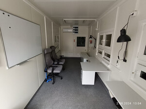 Wnętrze mobilnego kontenera biurowego. Od lewej na ścianie zamontowana biała tablica suchościeralna, na podłodze stoją dwa krzesła obrotowe. Po prawej stronie szerokie biurko a po lewej i prawej stronie na ścianie zamontowane na ruchomym ramieniu czarne kinkiety Wnętrze mobilnego kontenera biurowego. Od lewej na ścianie zamontowana biała tablica suchościeralna, na podłodze stoją dwa krzesła obrotowe. Po prawej stronie szerokie biurko a po lewej i prawej stronie na ścianie zamontowane na ruchomym ramieniu czarne kinkiety