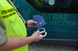 Funkcjonariusz w lewej ręce trzyma cztery paszporty, w prawej kajdanki, w tle znajduje się samochód Straży Granicznej. Funkcjonariusz w lewej ręce trzyma cztery paszporty, w prawej kajdanki, w tle znajduje się samochód Straży Granicznej.