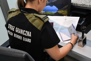  Ujęcie z tyłu siedzącej przy biurku funkcjonariuszki Straży Granicznej. W prawej dłoni trzyma paszport. Obok na blacie stoi stempel. Na koszulce napis: Straż Graniczna Polish Border Guard.