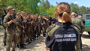  Funkcjonariuszka Straży Granicznej, na plecach napis Straż Graniczna Polish Border Guard. Z lewej strony ustawieni uczniowie z klas o profilu wojskowym i policyjnym