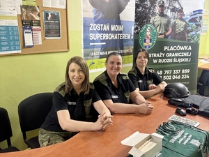  Trzy uśmiechnięte funkcjonariuszki Straży Granicznej siedzą przy stole z artykułami reklamowymi. W tle baner SG i plakat z napisem &quot;Zostań moim superbohaterem&quot;!