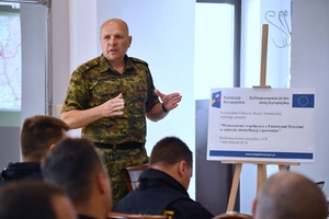  Prowadzący spotkanie. Po prawej stronie na sztaludze plakat z informacjami: Fundusze Europejskie. Dofinansowane przez Unię Europejska. Komendant Główny Straży Granicznej realizuje projekt: „Wzmocnienie współpracy z Państwami trzecimi w zakresie identyfikacji i powrotów”. Dofinansowanie projektu z UE: 7 milionów zł.