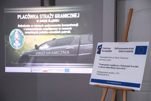  Na ścianie wyświetlony slajd z napisem: Placówka Straży Granicznej w Rudzie Śląskiej. Szkolenie w ramach podnoszenia kompetencji i wzmocnienia współpracy w ramach wspólnych polsko-czeskich patroli. Po prawej stronie na sztaludze plakat z informacjami: Fundusze Europejskie. Dofinansowane przez Unię Europejska. Komendant Główny Straży Granicznej realizuje projekt: „Wzmocnienie współpracy z Państwami trzecimi w zakresie identyfikacji i powrotów”. Dofinansowanie projektu z UE: 7 milionów zł.